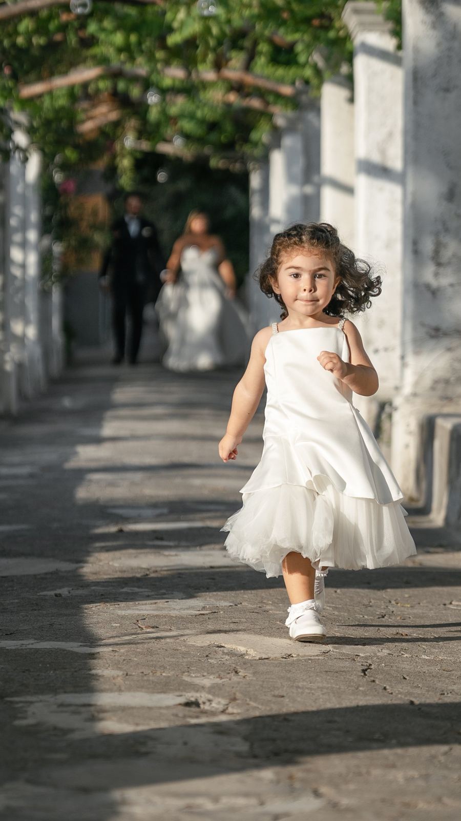 Una bambina vestita di bianco corre sotto il pergolato, gli sposi alle sue spalle
