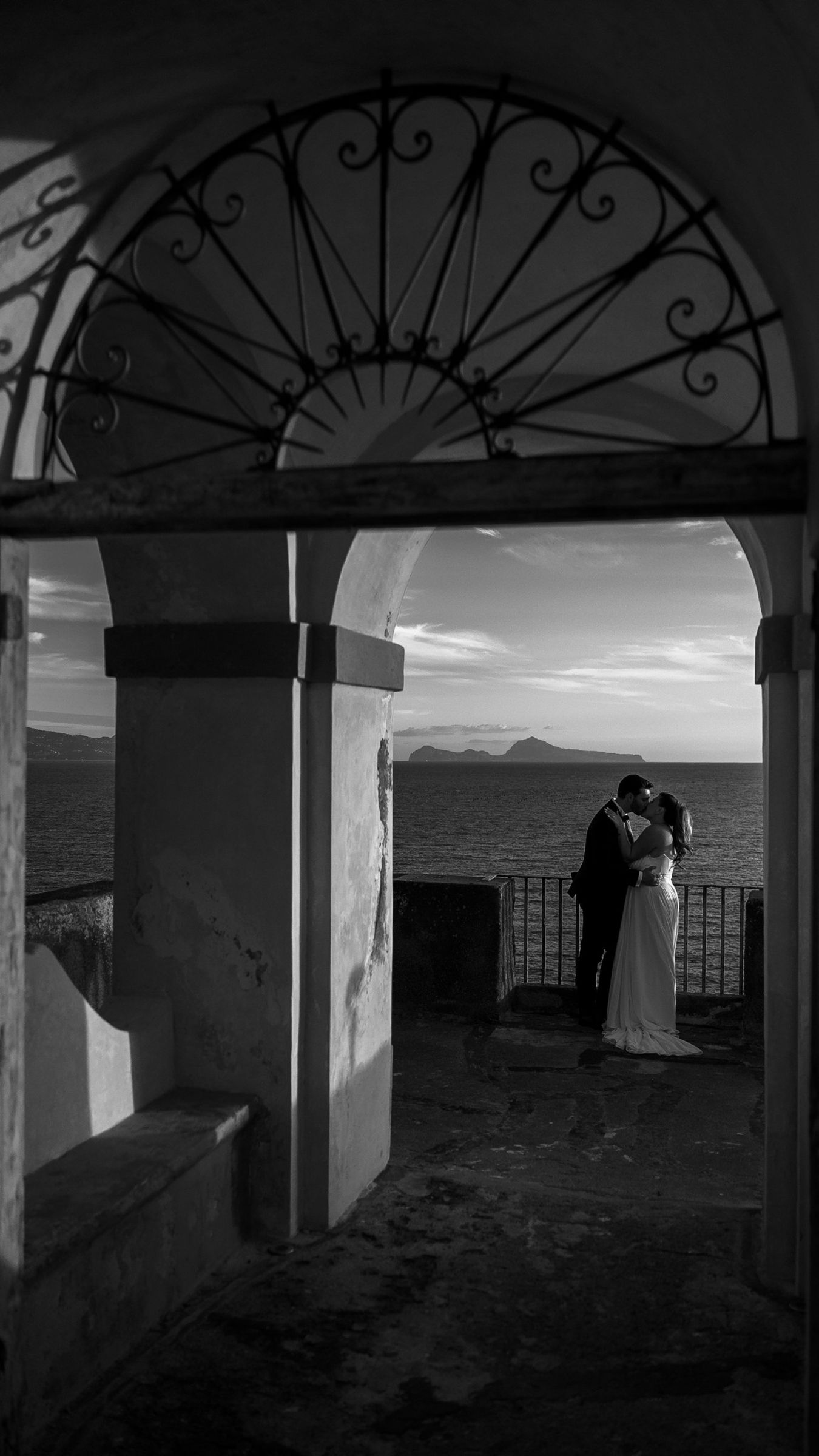 Gli sposi si baciano sotto un arco affacciato sul mare, con Capri all'orizzonte, in bianco e nero