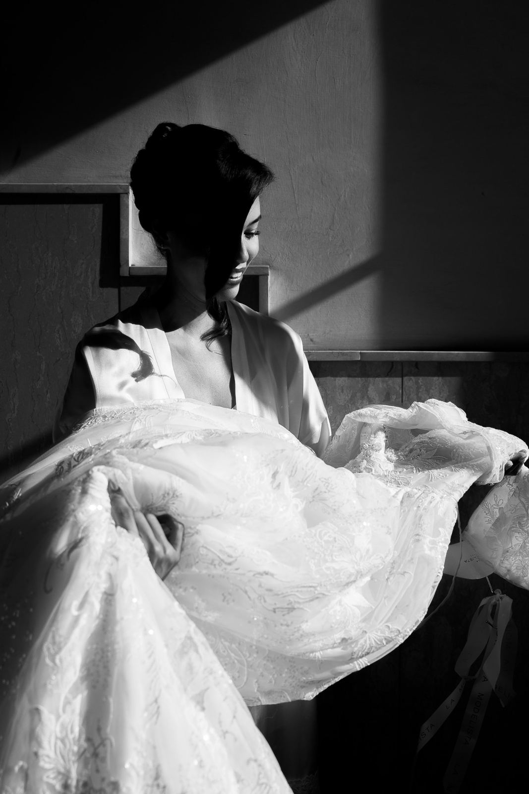 La sposa in vestaglia solleva l'abito in una lama di luce, in bianco e nero