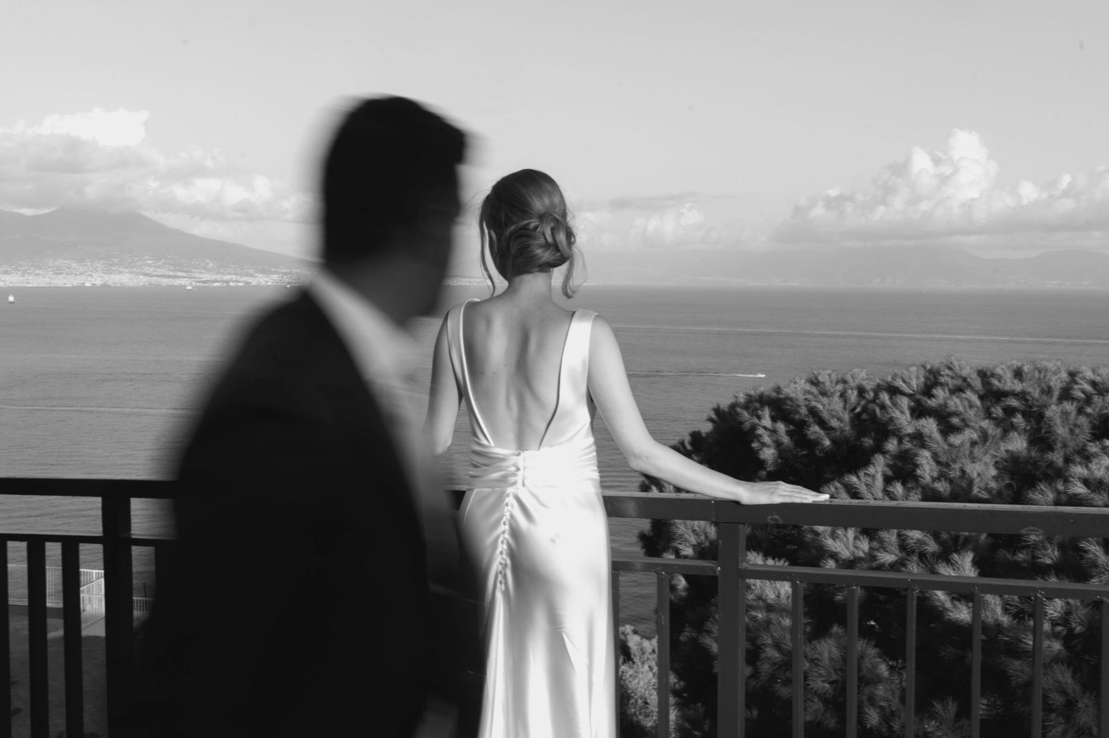 La sposa sul balcone guarda il golfo, lo sposo passa mosso in primo piano, in bianco e nero