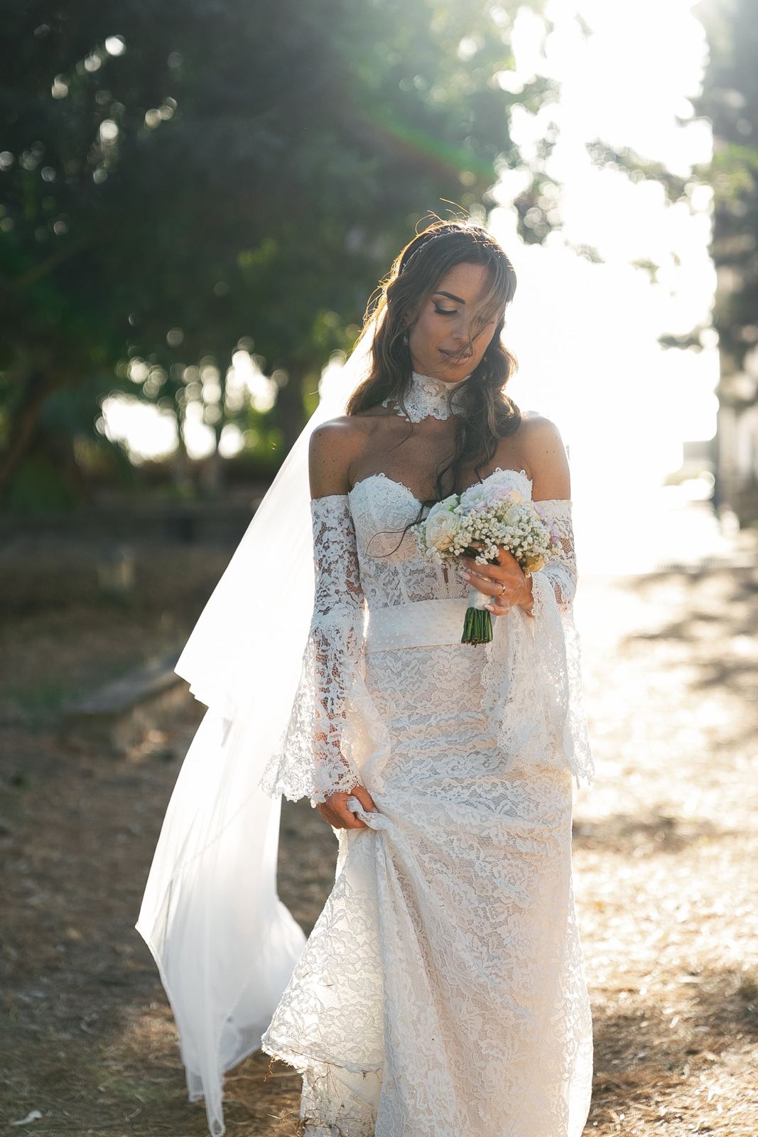La sposa cammina controluce con l'abito di pizzo e il bouquet